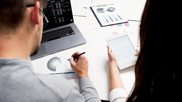 Two business professionals analyzing financial reports using a laptop and tablet in a modern office setting.